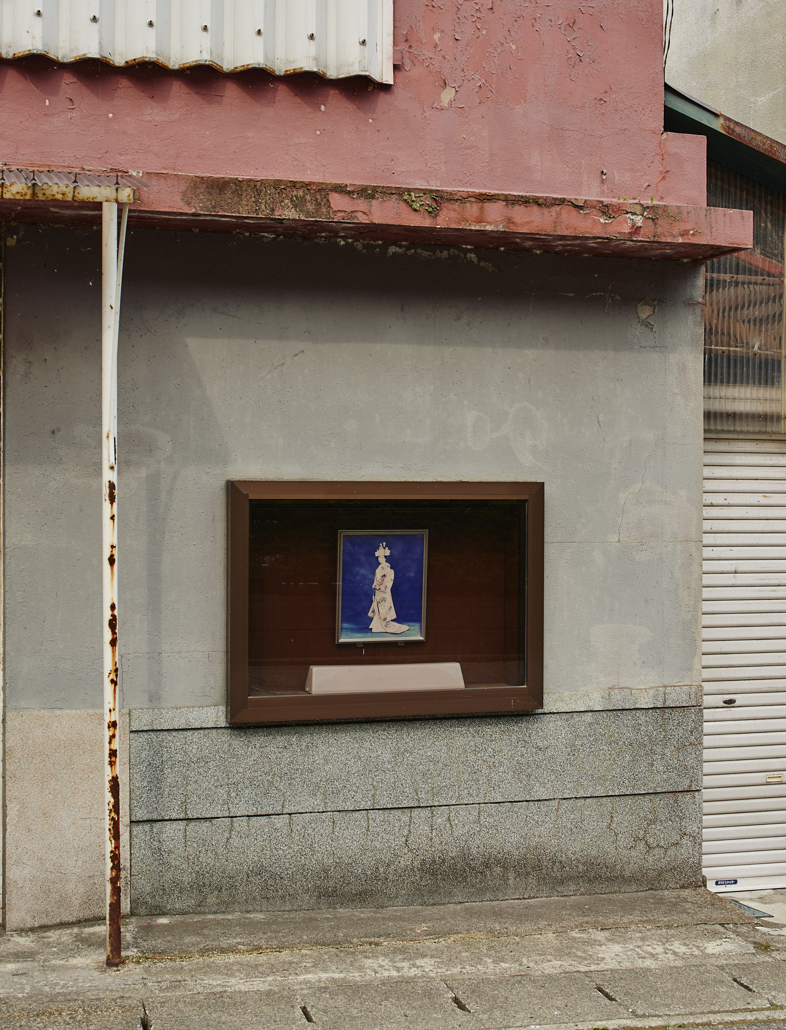 Framed artwork of a woman in traditional clothing displayed on the exterior of a weathered, pink-trimmed building with a roll-up door.