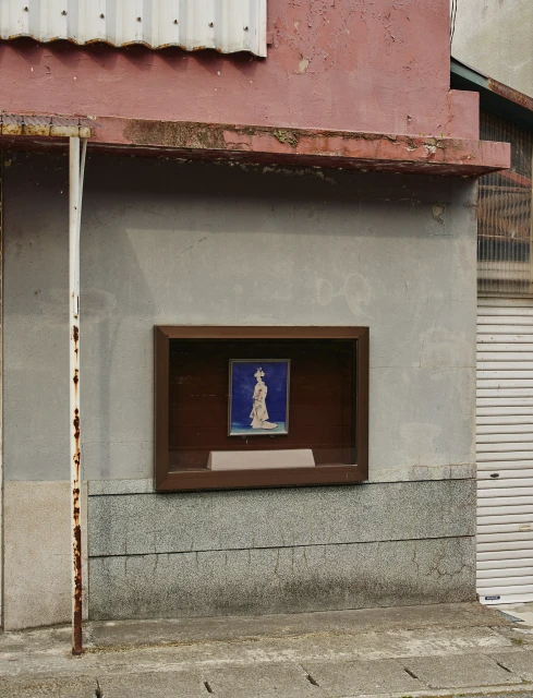Framed artwork of a woman in traditional clothing displayed on the exterior of a weathered, pink-trimmed building with a roll-up door.