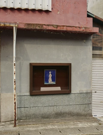Framed artwork of a woman in traditional clothing displayed on the exterior of a weathered, pink-trimmed building with a roll-up door.