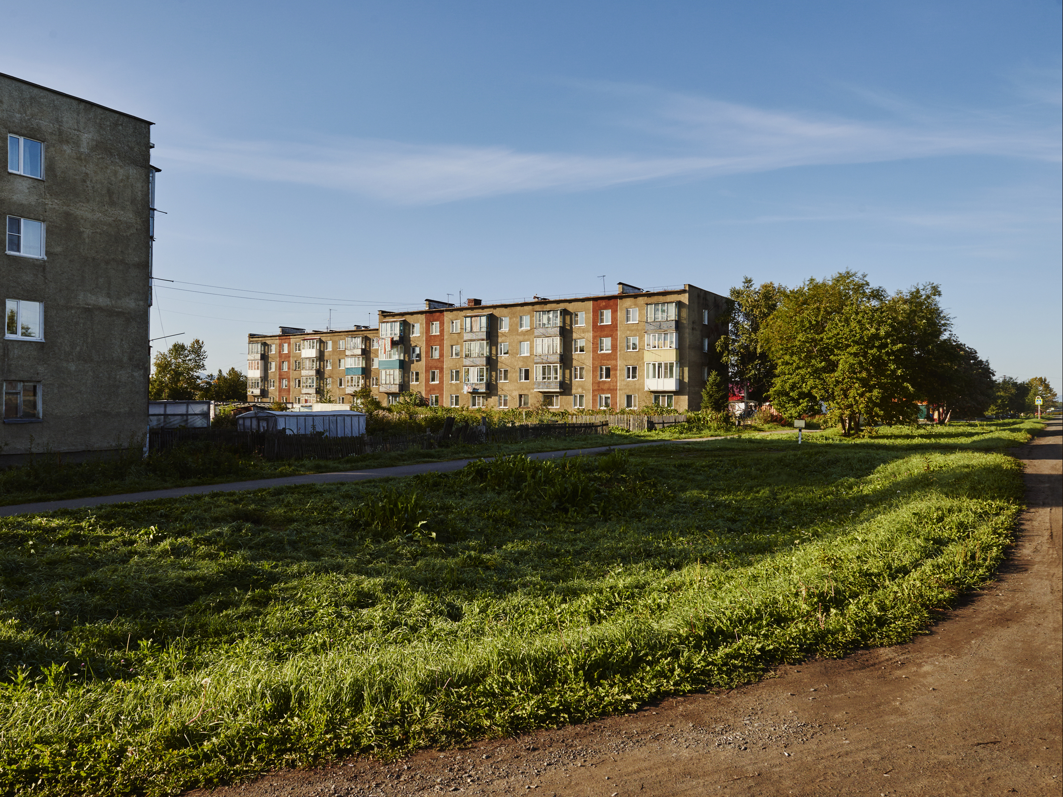 Residential buildings in a rural neighborhood with green grass, trees, and a dirt road under a blue sky on a bright, sunny day.