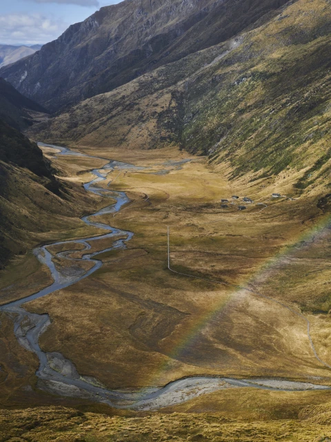 Panoramic view of a mountain valley with a winding river, brown grassland, distant buildings, and a faint rainbow under a blue sky.