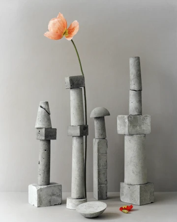 Still life shot of gray concrete geometric blocks arranged with a single peach-colored poppy flower in indoor setting with neutral backdrop.