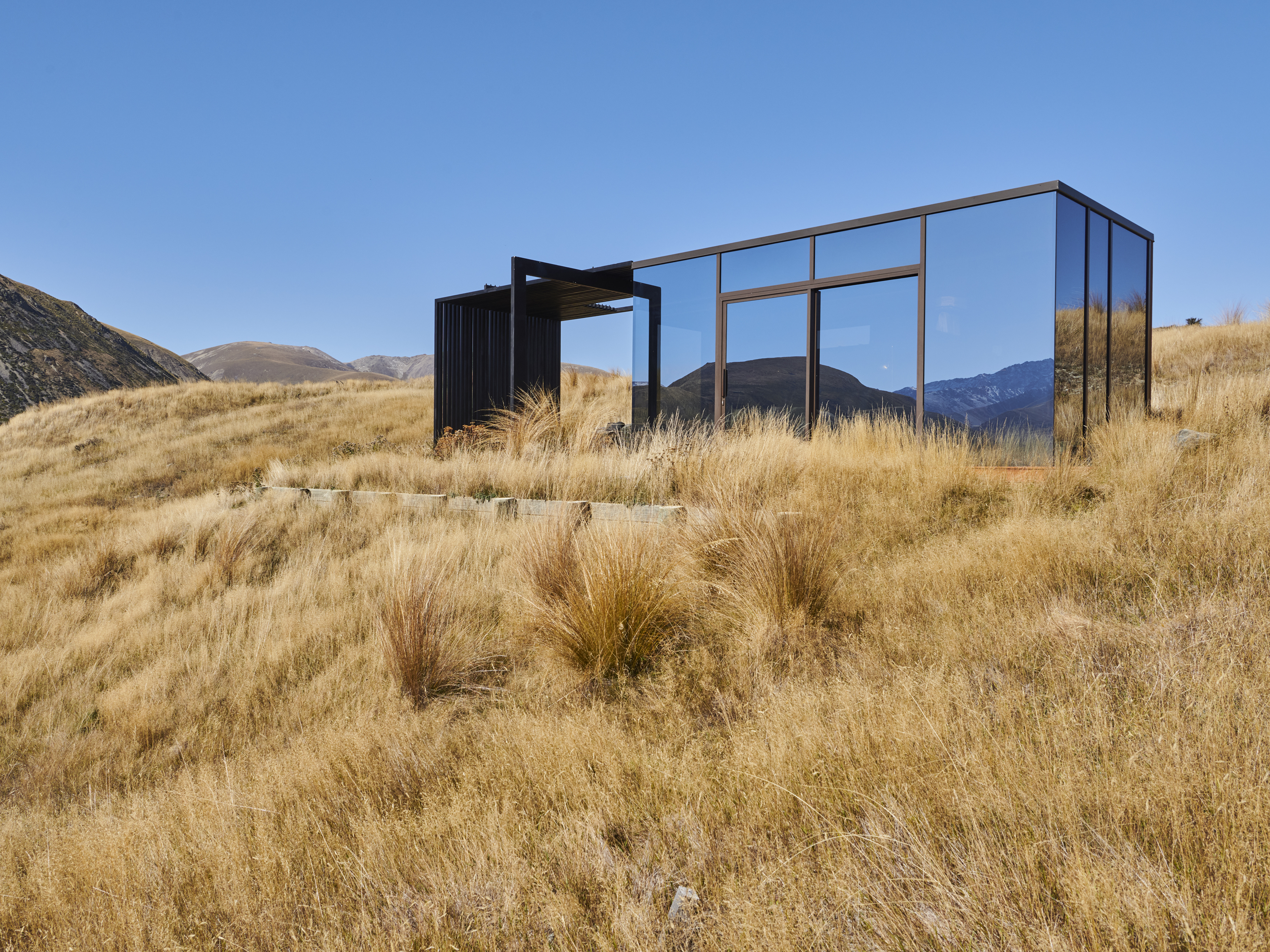 Modern mirrored house on a grassy hillside with mountains in the background, under a clear blue sky on a sunny day.