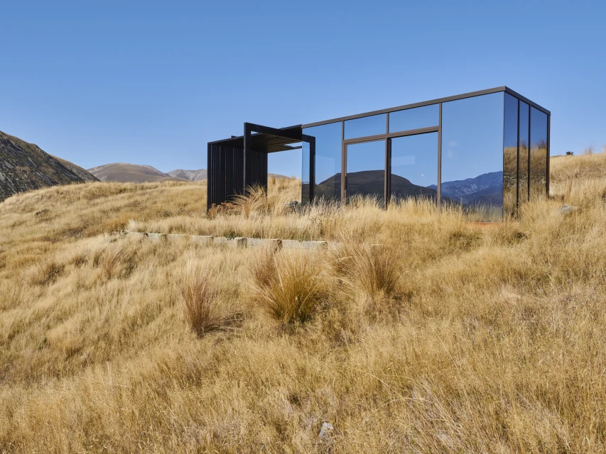 Modern mirrored house on a grassy hillside with mountains in the background, under a clear blue sky on a sunny day.