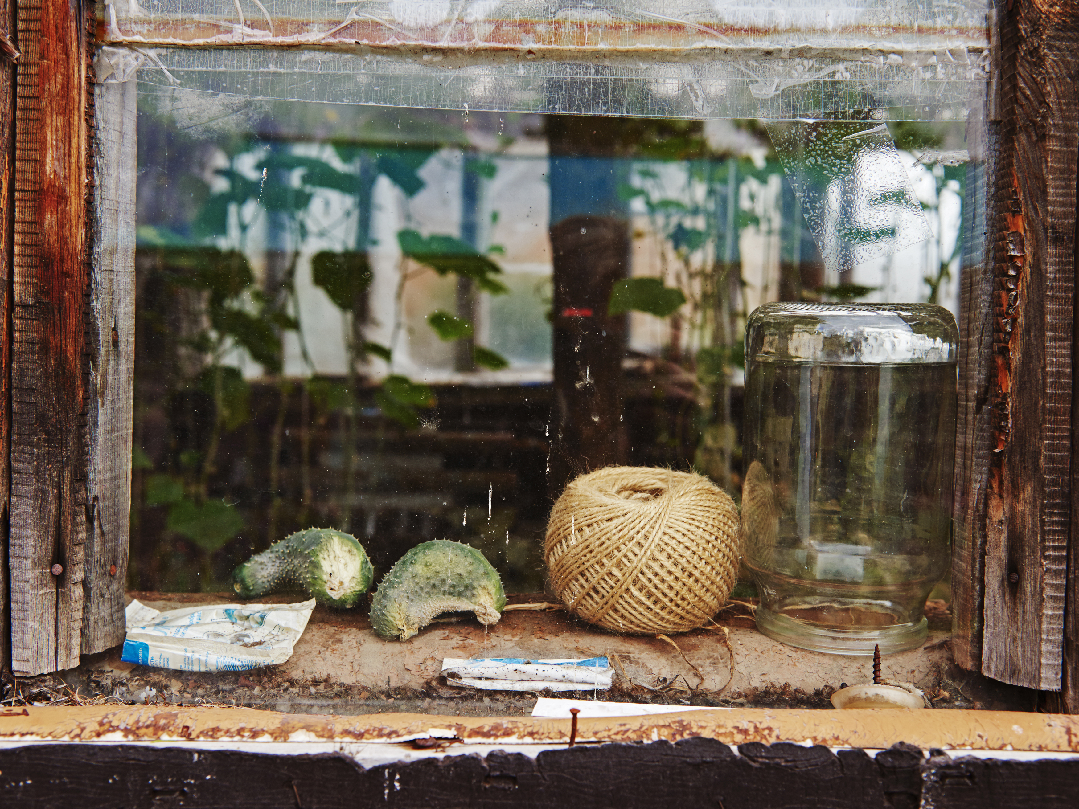 Still life on a windowsill featuring an empty jar, ball of twine, and prickly squash remnants, all viewed through an old wooden window.
