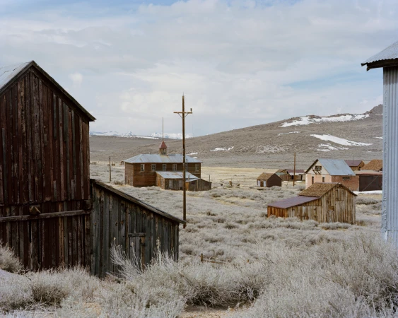 Historic wooden mining town buildings set in an arid landscape with distant snow-capped mountains under an overcast sky.