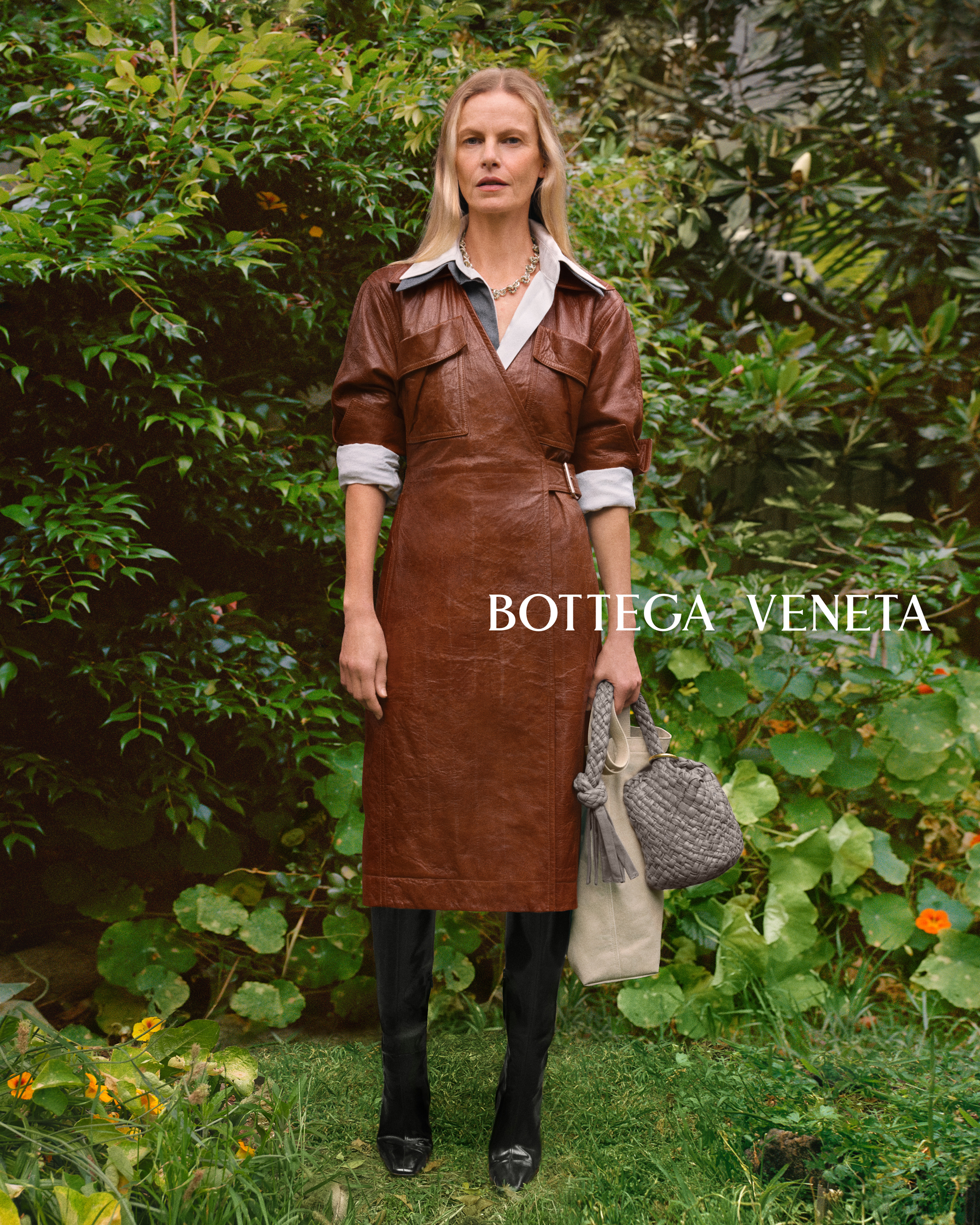 Fashion editorial shot of a blonde woman wearing a brown dress, black boots and holding a purse, posing outdoors with green foliage.