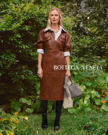 Fashion editorial shot of a blonde woman wearing a brown dress, black boots and holding a purse, posing outdoors with green foliage.