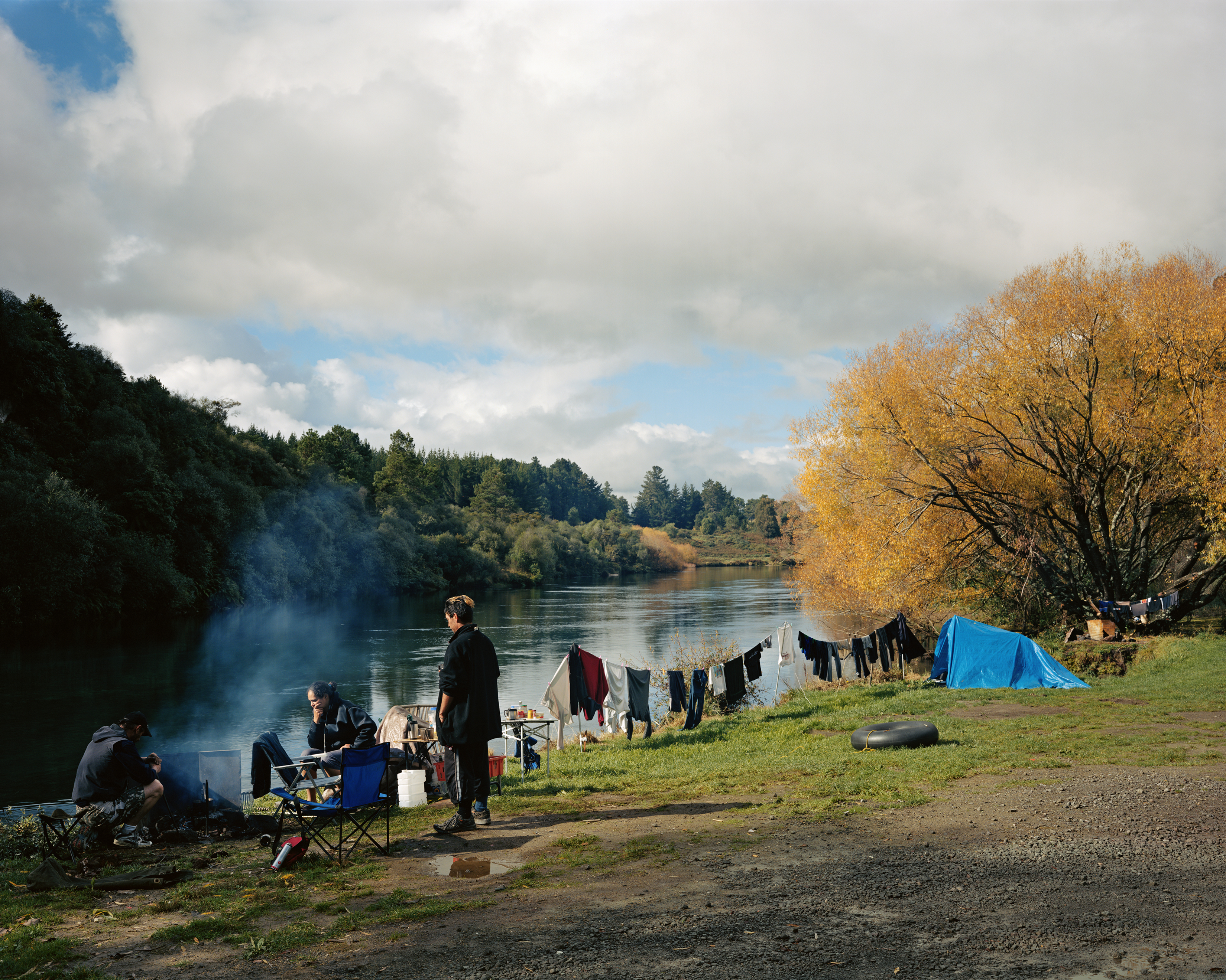 Outdoor camping scene by a river with three individuals near a fire, clothes hanging to dry, and a blue tent on the grassy bank.