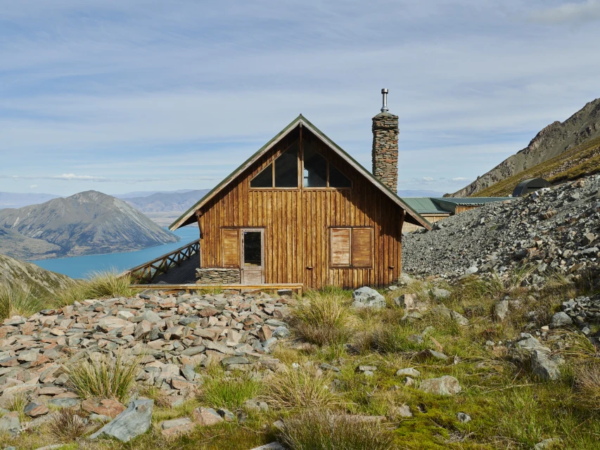Exterior view of a rustic wood cabin with a stone chimney on a rocky hill overlooking a turquoise lake and mountains under a blue sky.
