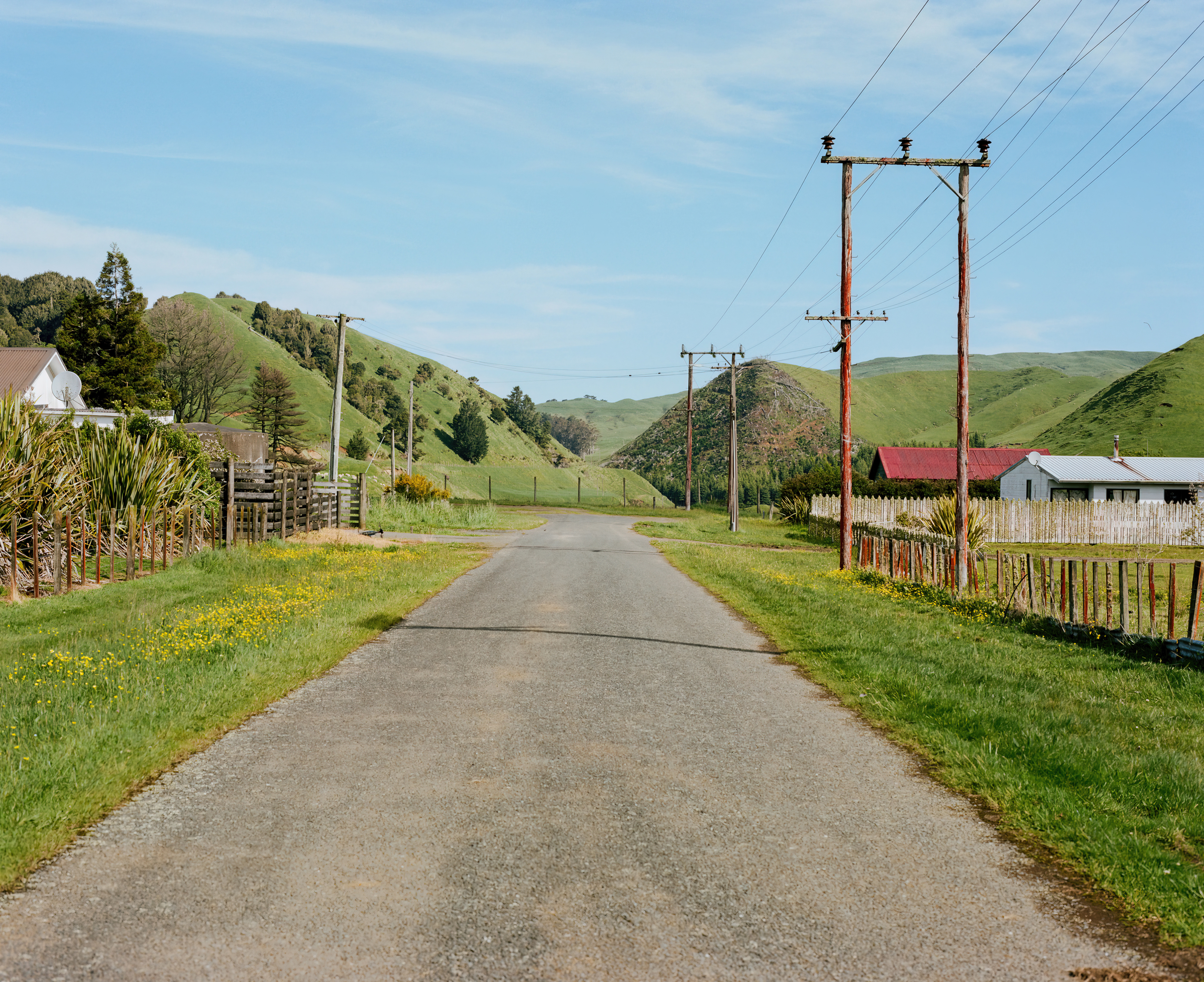 Gravel road leads through a rural landscape with green hills, vegetation, houses, and telephone poles under a clear blue sky.