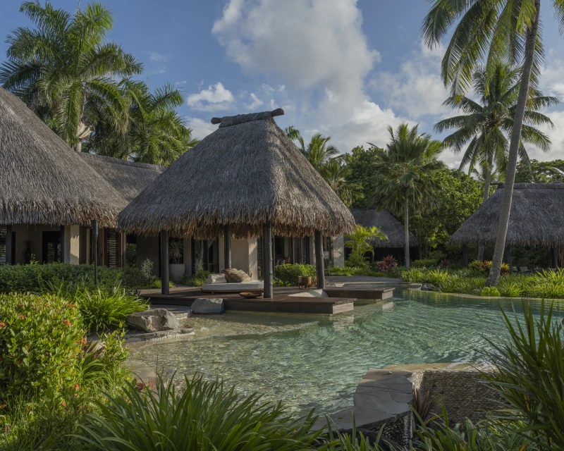 Luxurious tropical resort with thatched-roof bungalows, turquoise water, and lush vegetation under a partly cloudy sky. Vacation ambiance.