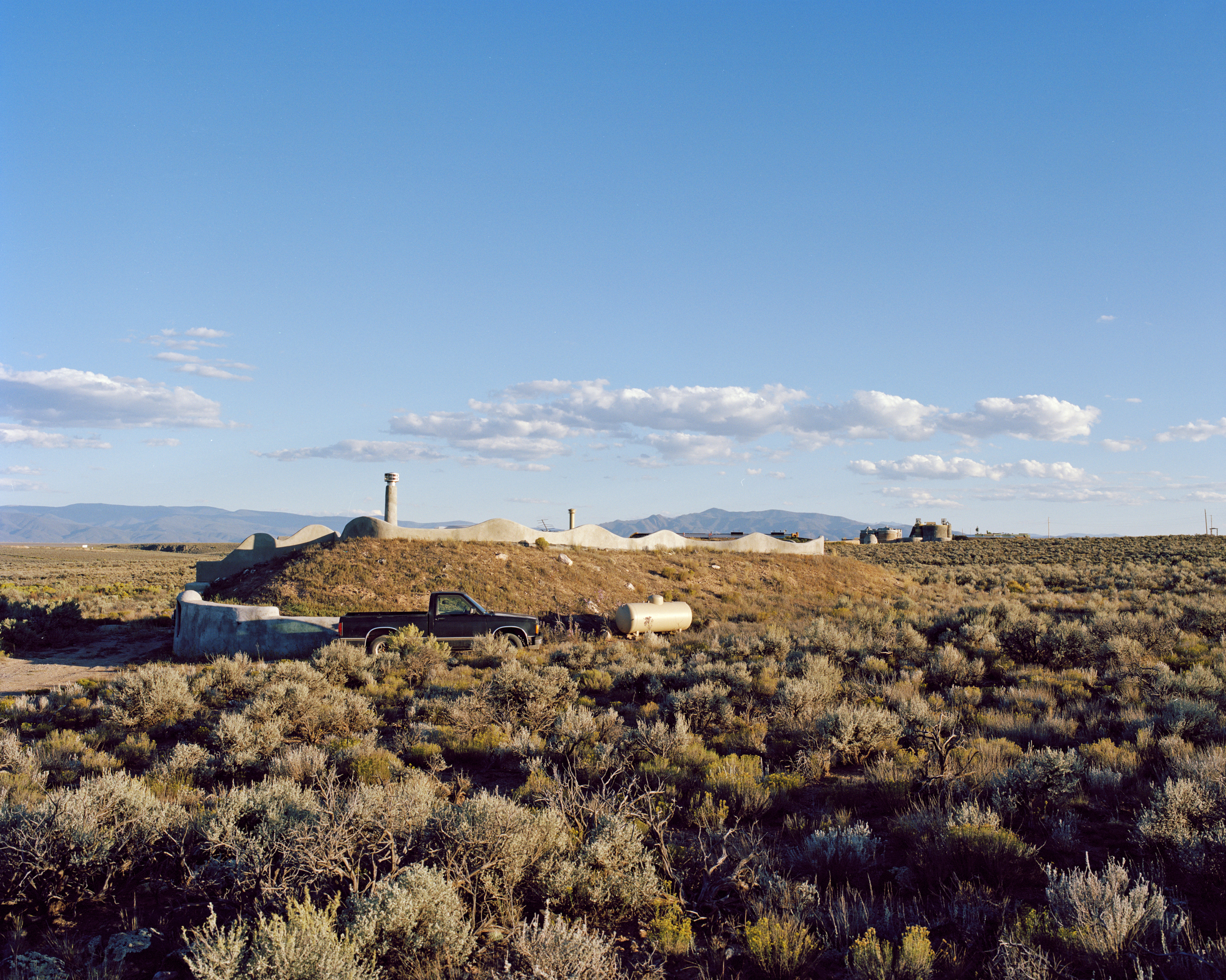 Exterior daytime shot of a black pick-up truck parked in front of an earth berm house in a rural, grassy field under a blue sky.