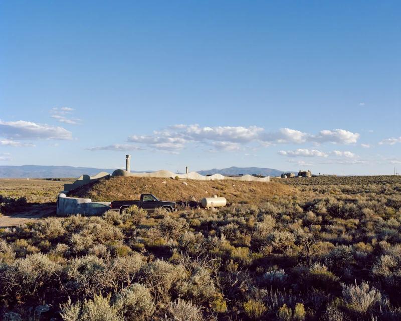 Exterior daytime shot of a black pick-up truck parked in front of an earth berm house in a rural, grassy field under a blue sky.