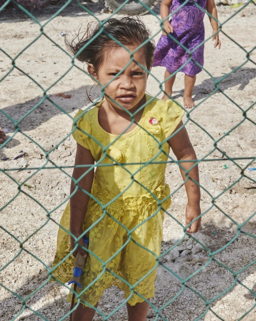 A young child in a yellow dress stands behind a green chain-link fence in an outdoor, sunlit area. Another child is visible in the background.
