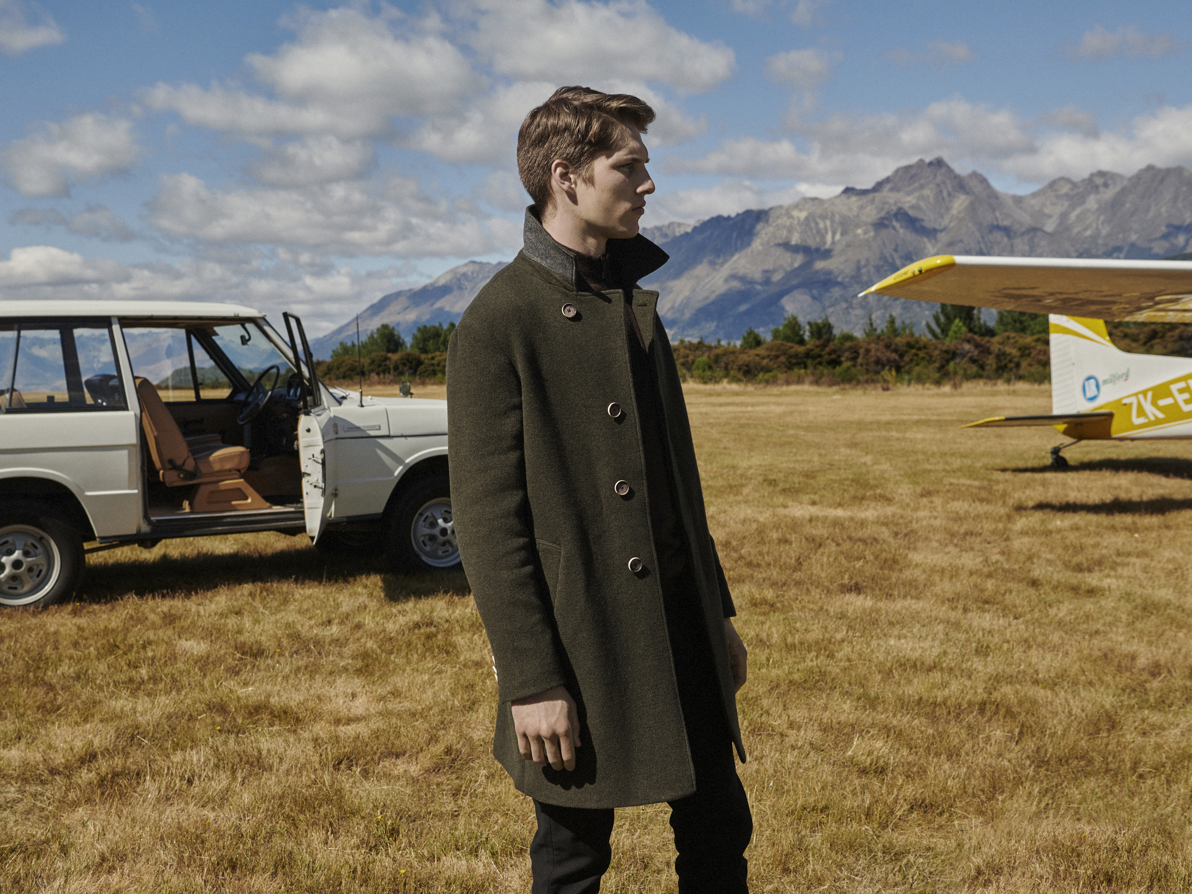 A fair-skinned adult male wearing a green coat stands in a grassy field with a Range Rover and small plane in the background.