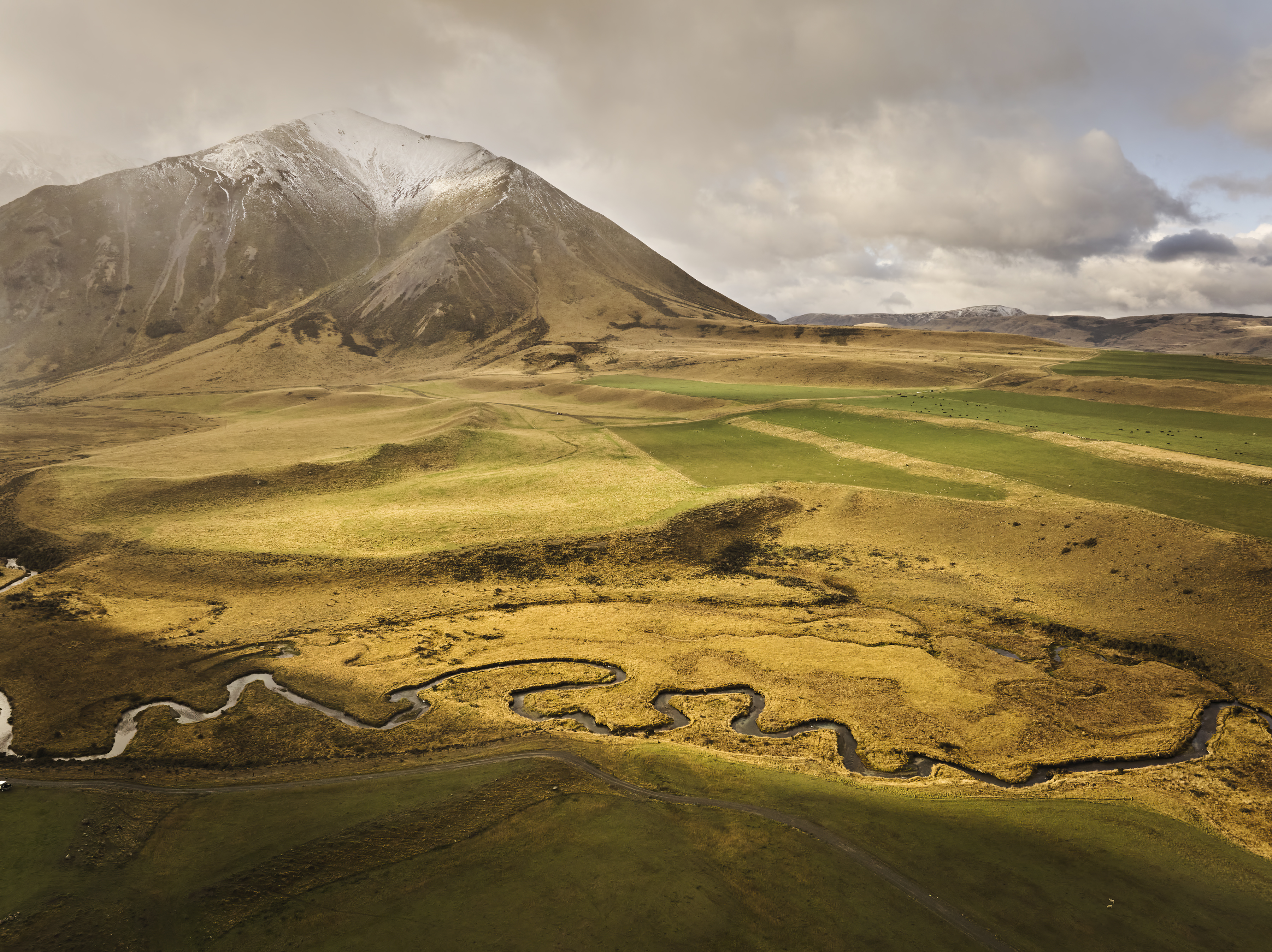 Scenic aerial shot of a winding river flowing through golden grassland toward a snow-capped mountain under a cloudy sky.