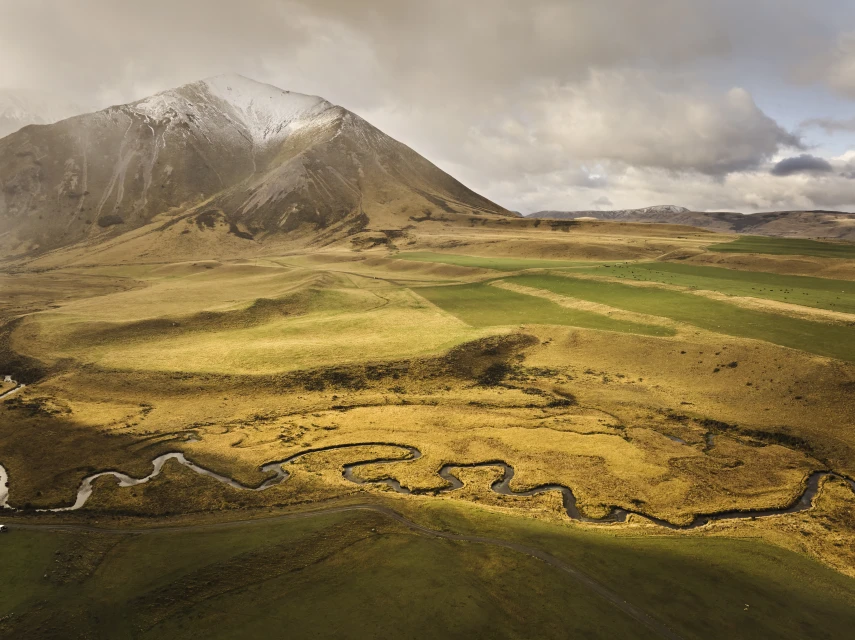 Scenic aerial shot of a winding river flowing through golden grassland toward a snow-capped mountain under a cloudy sky.