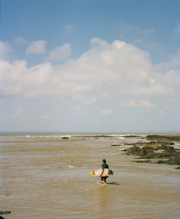 Surfer carrying a colourful surfboard walking through shallow coastal water under a bright sky with scattered clouds.