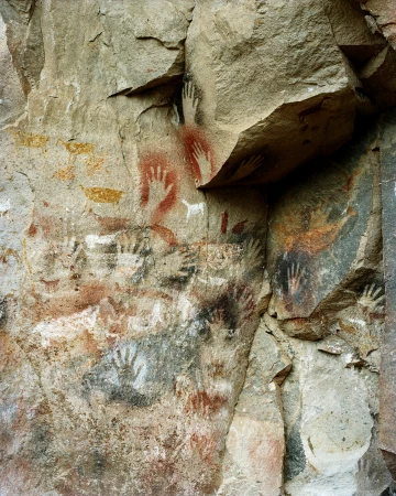 Rock wall with ancient hand paintings in red, white, black, and yellow pigment in a cave setting with rough stone texture.