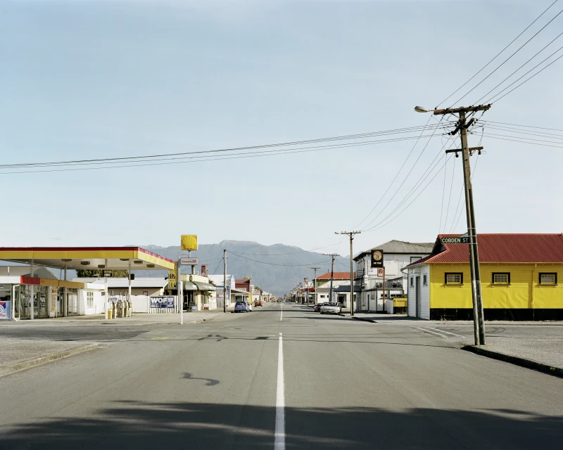 Street view in a small town featuring a road stretching into the distance, buildings, power lines, and a mountain range backdrop under a clear sky.