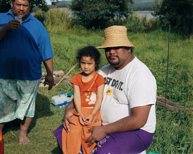 A father and daughter are sitting together in a grassy field with a man in the background standing next to a lake surrounded by trees.