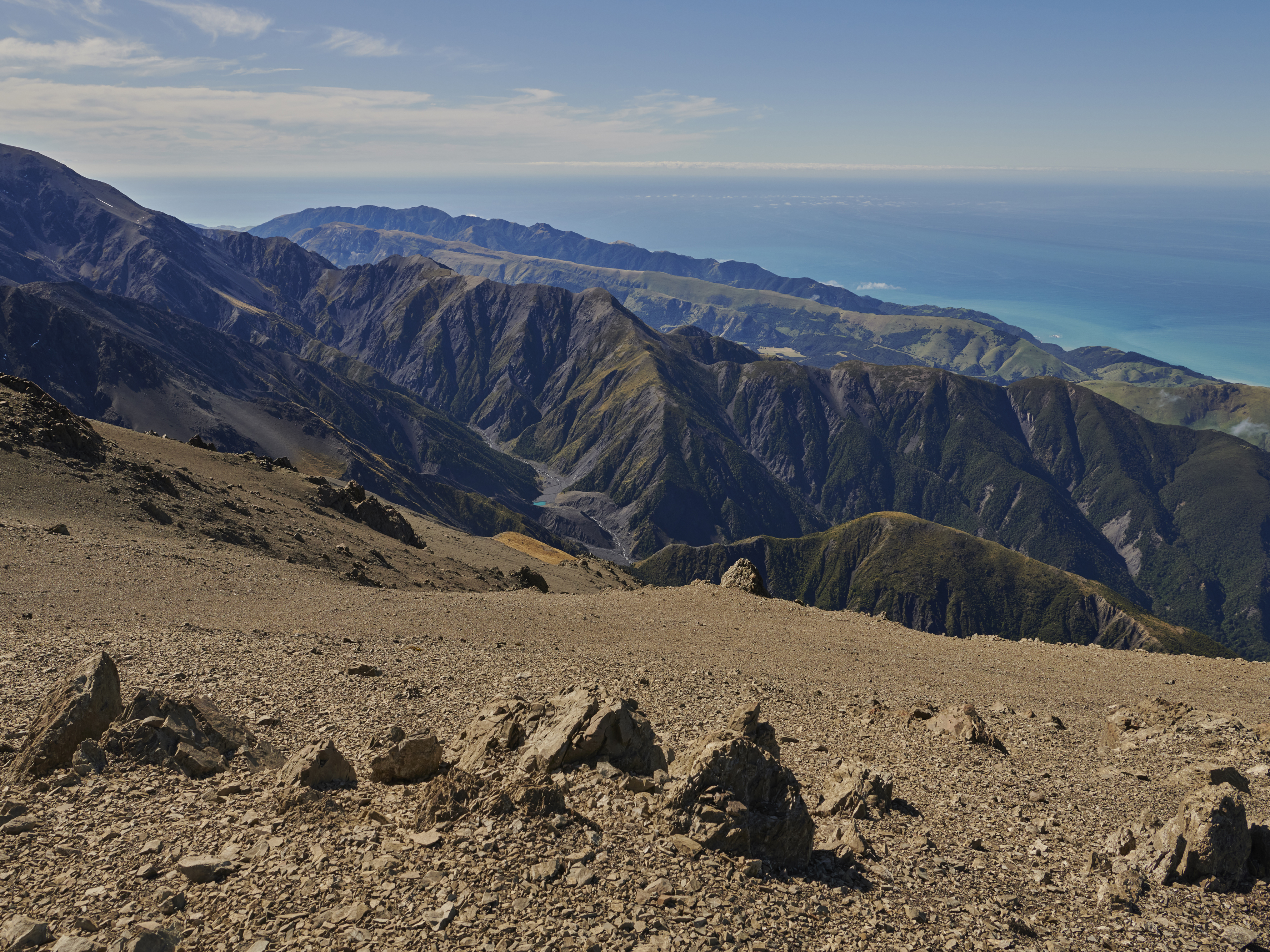 Landscape view of rocky mountain slope overlooking valleys, forested hills, and a blue ocean under a clear sky, creating a scenic vista.