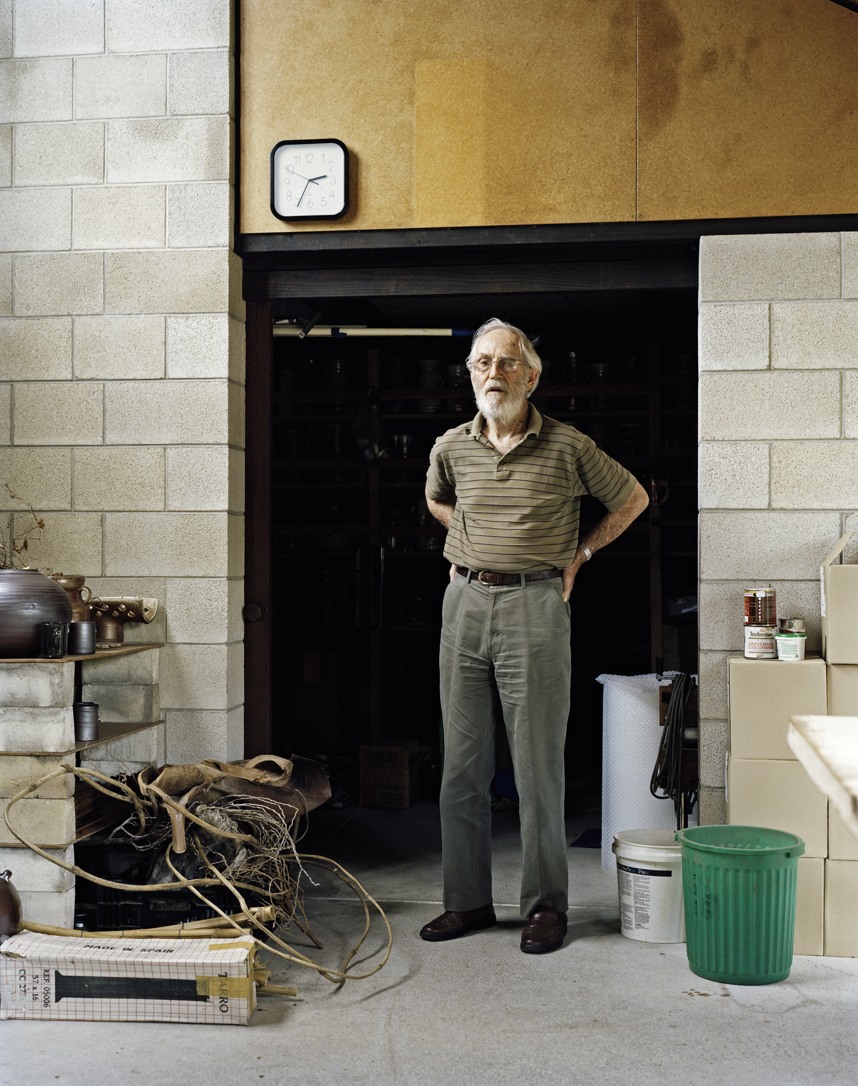 Full-length portrait of an elderly man with glasses, wearing a striped shirt, in front of a dark doorway in an industrial-style indoor setting.