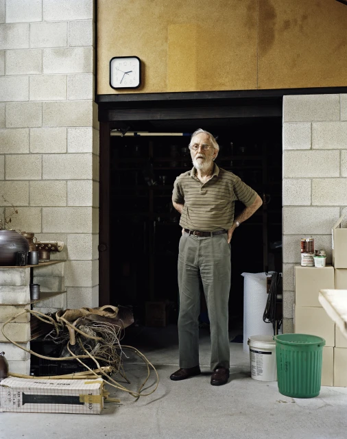 Full-length portrait of an elderly man with glasses, wearing a striped shirt, in front of a dark doorway in an industrial-style indoor setting.