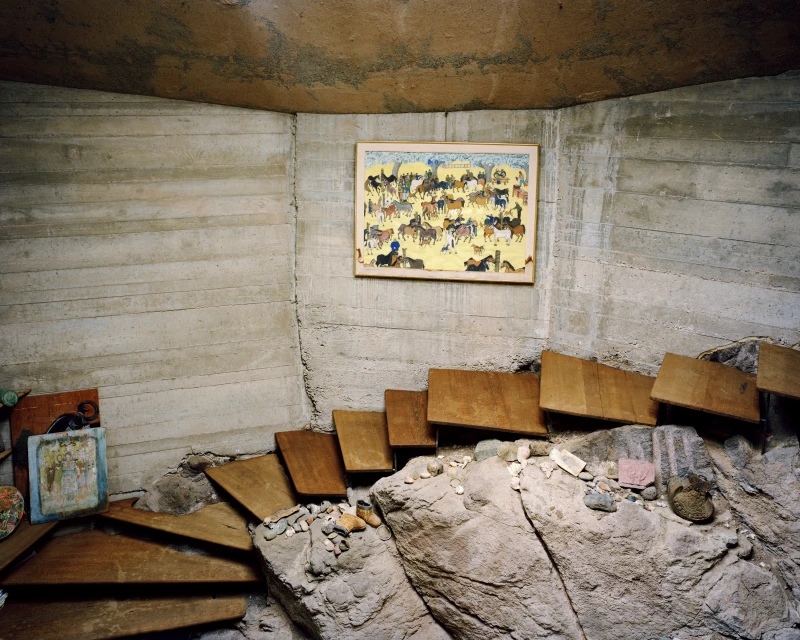 Interior view with a framed painting of horses hanging on a concrete wall above wooden steps and a stone base in a room.