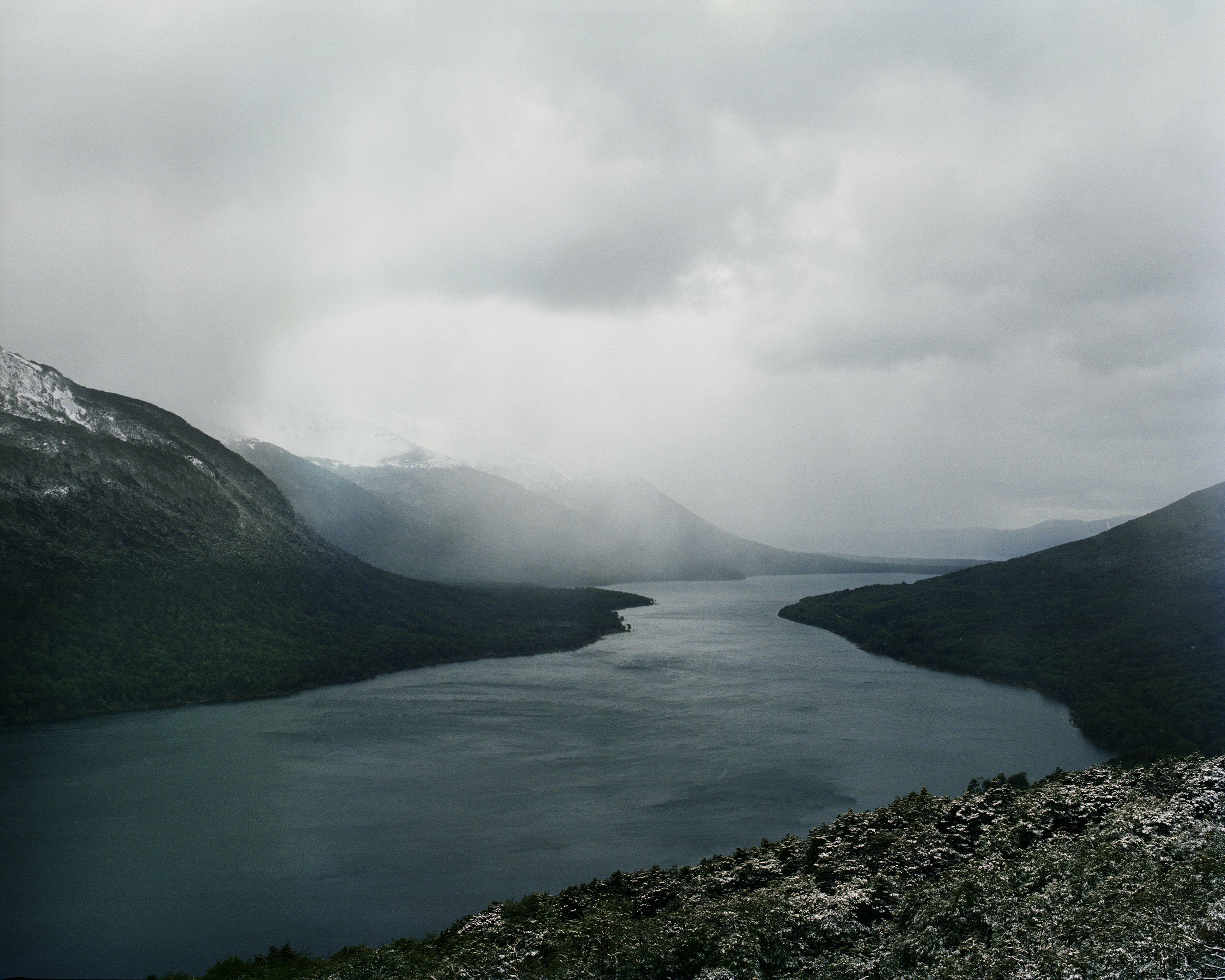 Overhead landscape view of a large lake surrounded by green mountains under a cloudy sky, with snow on the mountain peaks.