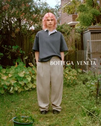 A person with pink hair wearing a dark polo shirt and beige trousers stands in a lush garden by Derek Henderson.