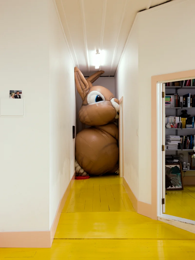 Giant inflatable brown rabbit wedged at the end of a narrow hallway with bright yellow wooden floors.