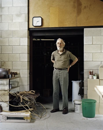 Elderly man standing in an industrial workspace wearing a striped polo shirt and grey trousers by Derek Henderson