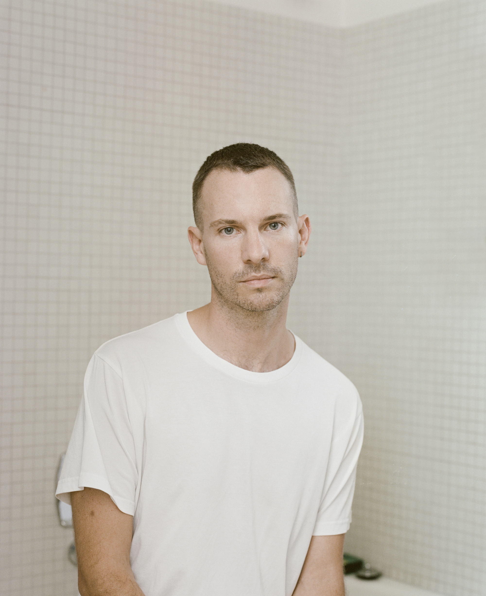Portrait of a man in a white t shirt standing in front of a tiled wall by photographer Derek Henderson