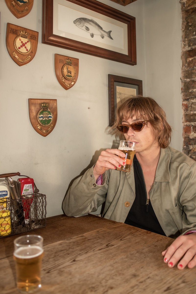 A fair-skinned person with brown hair and sunglasses drinks from a beer glass at a wooden table in an indoor pub setting.