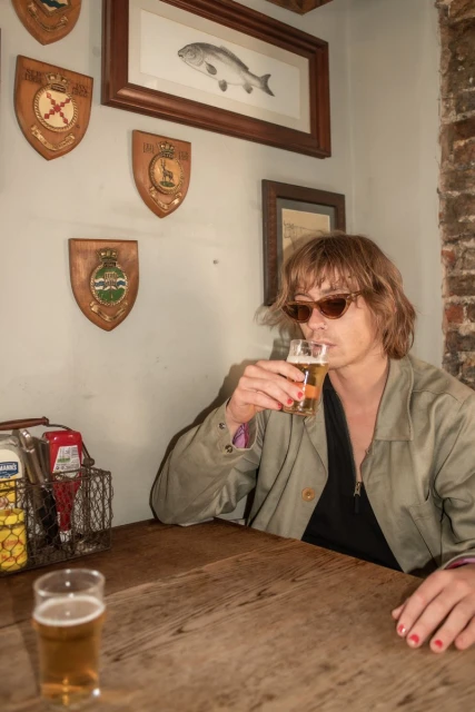 A fair-skinned person with brown hair and sunglasses drinks from a beer glass at a wooden table in an indoor pub setting.