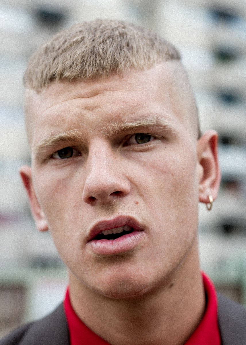 Close-up portrait of a young adult male with a unique haircut and neutral expression, wearing a red shirt and grey jacket, against a blurred urban background.