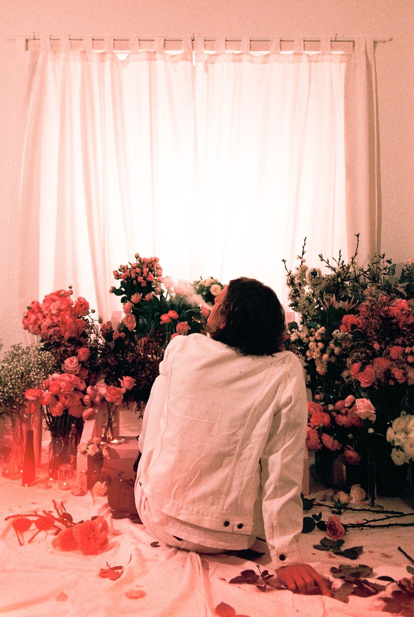 Woman in a white jacket sits surrounded by a vibrant display of roses and other flowers indoors with a soft, diffused light from a window.