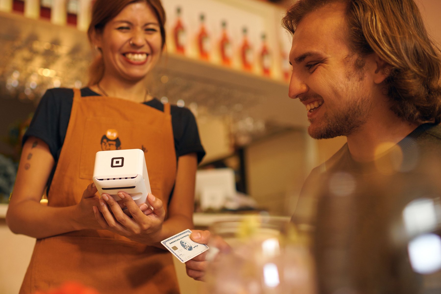 Adult female cashier hands card reader to adult male customer inside an indoor cafe environment with natural lighting and fair skin.