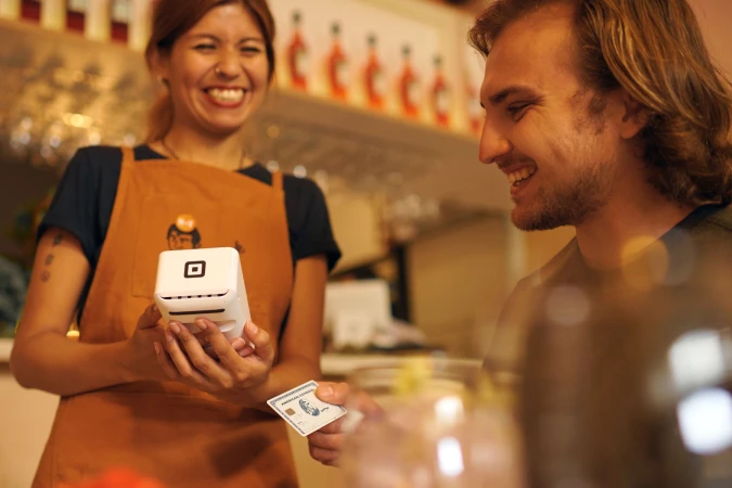 Adult female cashier hands card reader to adult male customer inside an indoor cafe environment with natural lighting and fair skin.