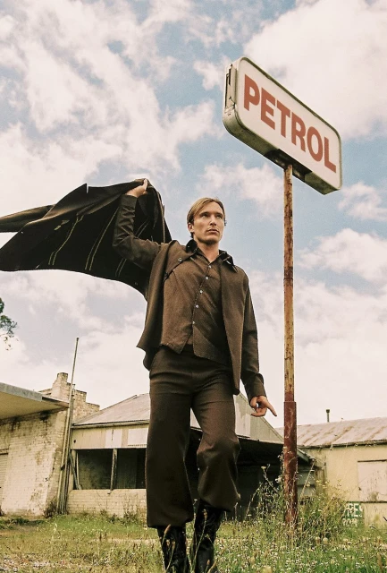 Adult male model in brown suit and cape stands under a vintage Petrol sign in a field of grass with old buildings and cloudy sky.