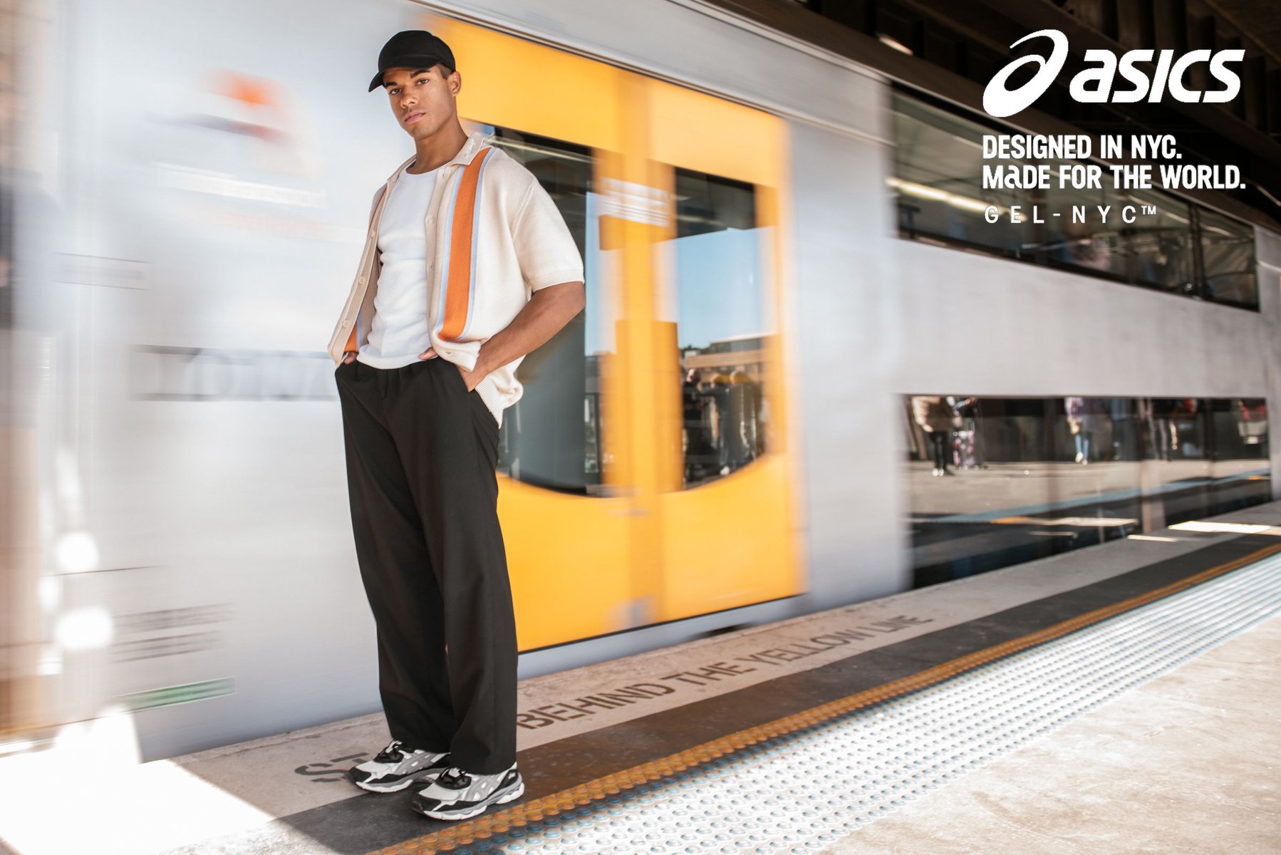 Man in casual fashion stands on a train platform with blurred train in the background, showcasing apparel and Asics sneakers.