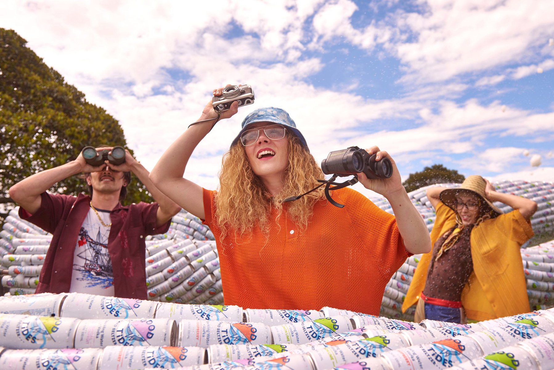 Three adults stand amidst a field of seltzer cans on a sunny day. They are looking through binoculars and a toy camera, smiling outdoors.