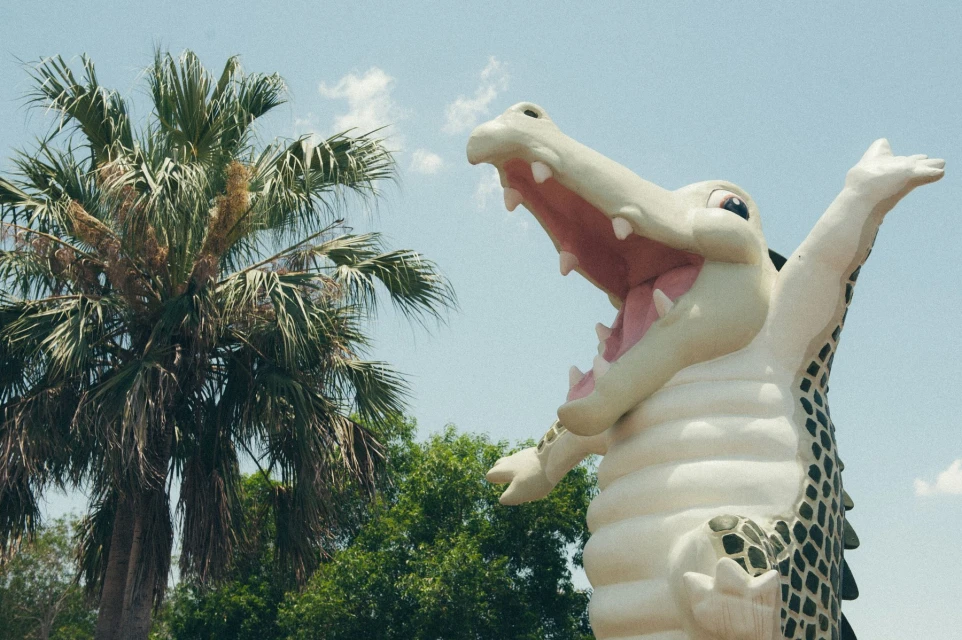 Large outdoor alligator sculpture with open mouth next to palm tree against blue sky in sunny weather; travel and tourism landmark.