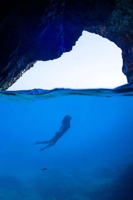 Woman swims in blue ocean water towards open sea cave with dramatic light. Underwater view of marine environment.