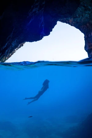 Woman swims in blue ocean water towards open sea cave with dramatic light. Underwater view of marine environment.