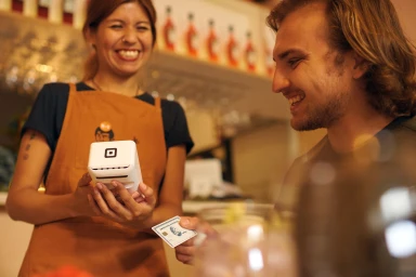 Smiling woman wearing a brown apron holding a small white payment terminal while a male customer pays with a credit card.