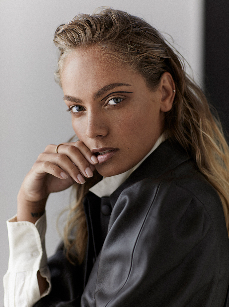 Portrait of a blonde woman with strong eyeliner and a black jacket, posing indoors against a neutral background, shot in soft natural light.