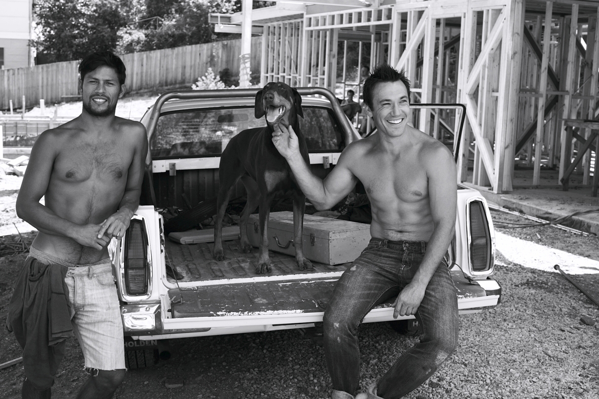 Two shirtless men pose with a dog in the back of a pickup truck at a construction site. Black and white shot, casual lifestyle vibe.
